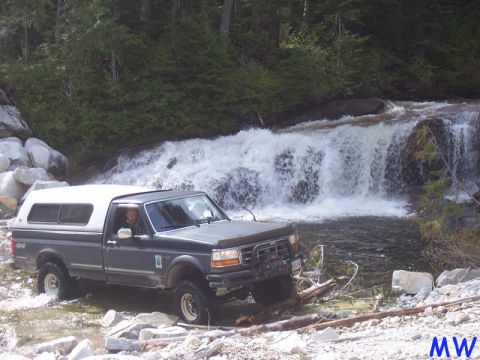 Dark gray Ford F-series pickup truck with white camper shell driving through river near waterfall.