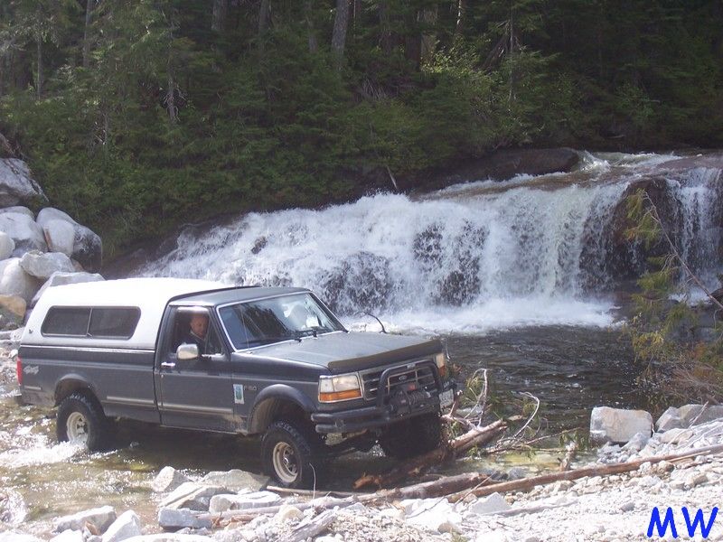 Dark gray Ford F-series pickup truck with white camper shell driving through river near waterfall.