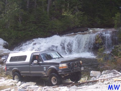 Dark gray Ford pickup truck with white camper shell driving through a stream near a waterfall.