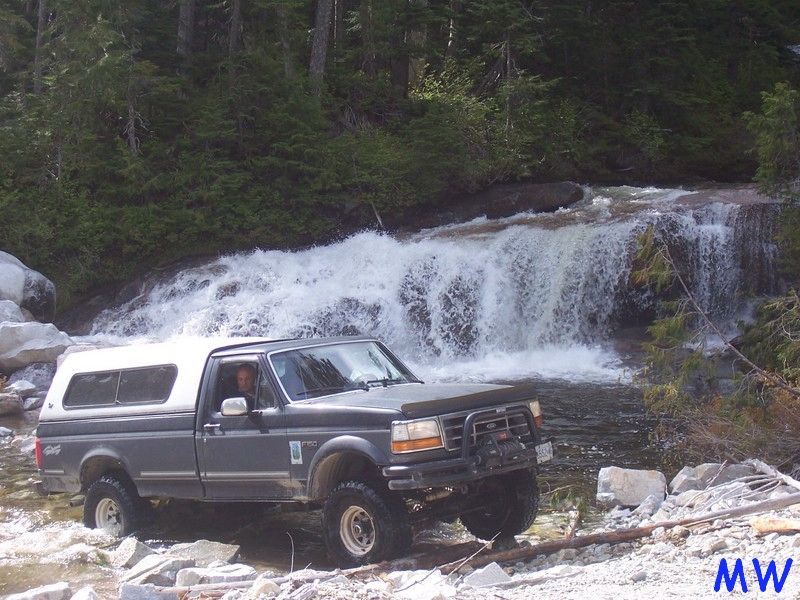 Dark gray Ford pickup truck with white camper shell driving through a stream near a waterfall.