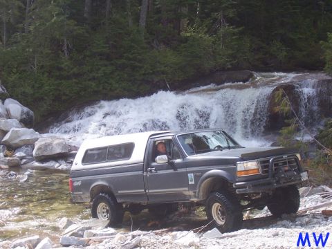 Dark gray pickup truck with white camper shell driving through a stream near a waterfall.