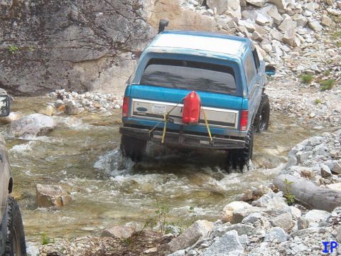 Blue 1985 Ford Bronco driving through a rocky stream with a red gas can on the back.