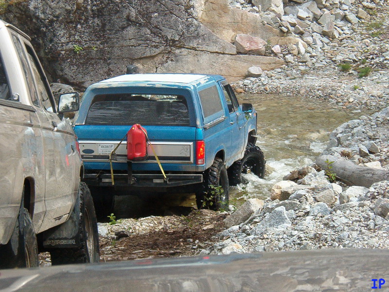 Blue Ford Bronco driving through a rocky stream, red gas can on rear.