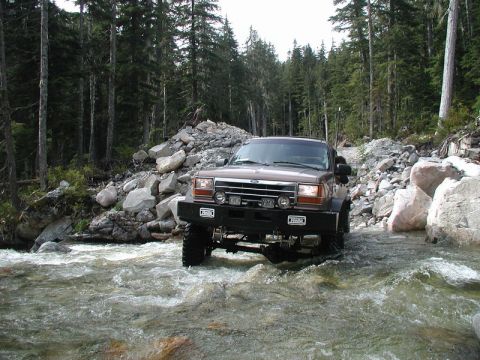 Lifted brown Ford Explorer driving through a river in a forest.