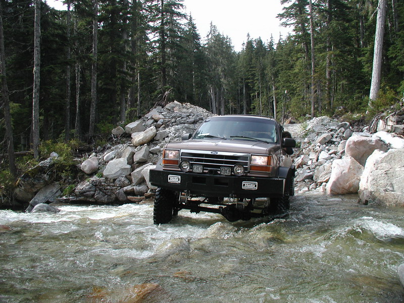 Lifted brown Ford Explorer driving through a river in a forest.