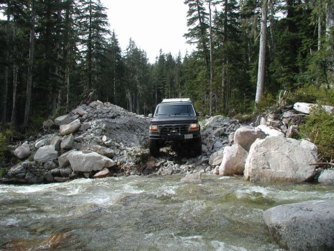 Dark SUV crossing a rocky river in a forest.
