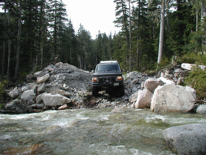 Dark SUV crossing a rocky river in a forest.