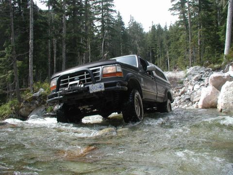 Dark Ford Bronco driving through a river in a forest