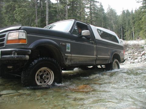 Dark Ford F-150 pickup truck with white camper shell driving through a stream in a forest.