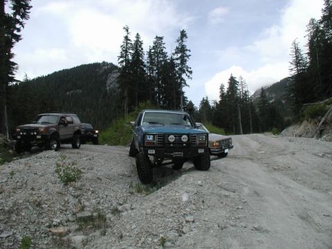 Three off-road vehicles on a mountain road: blue pickup, tan pickup, and brown SUV.