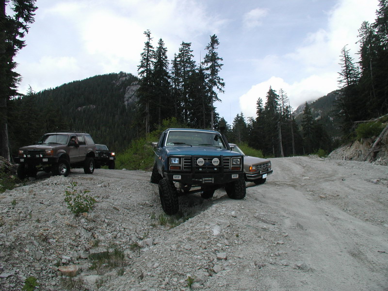 Three off-road vehicles on a mountain road: blue pickup, tan pickup, and brown SUV.