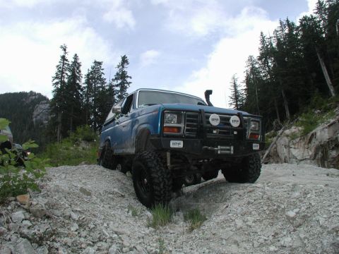 Blue 1980s Ford Bronco off-roading on a rocky hill with trees in the background.