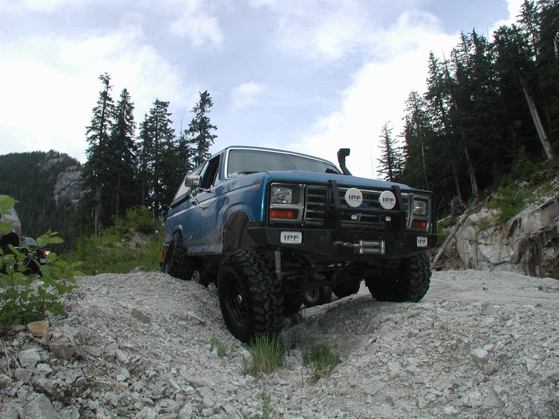 Blue 1980s Ford Bronco off-roading on a rocky hill with trees in the background.