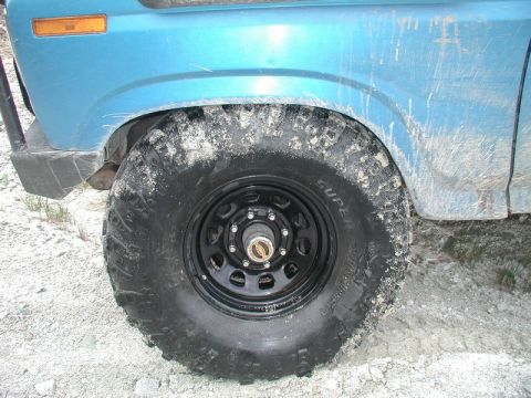 Blue 1985 Ford Bronco front wheel and fender with off-road tire covered in mud.
