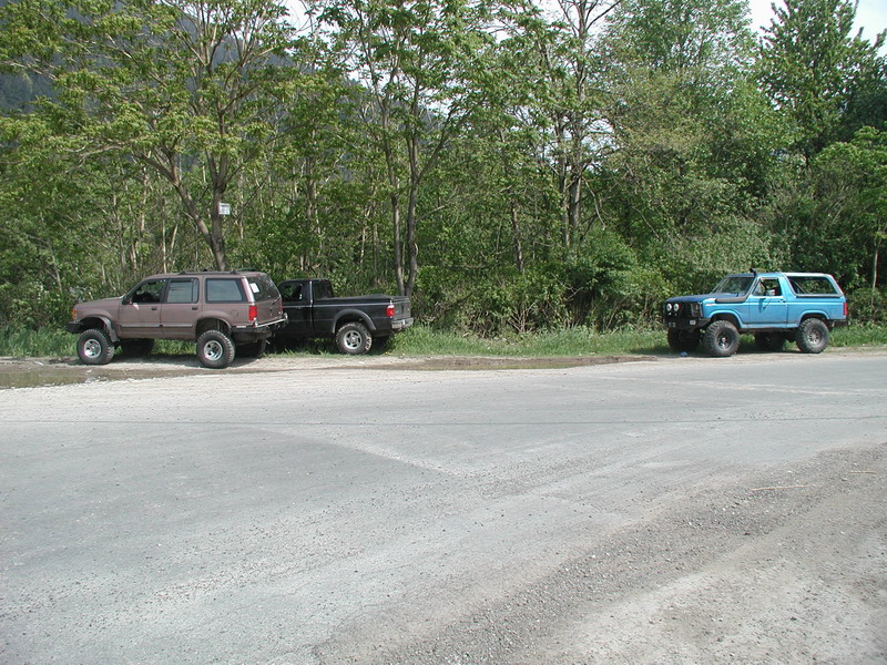 Lifted blue 1985 Ford Bronco parked on dirt road beside trees, with other vehicles in background.