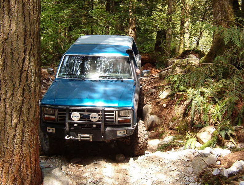 Blue 1980s Ford Bronco off-roading on a rocky forest trail.