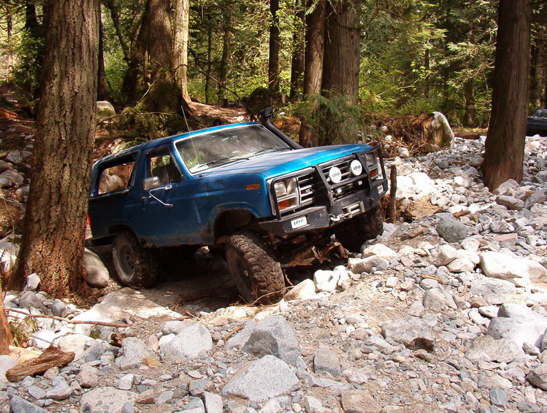 Blue 1980s Ford Bronco off-roading on a rocky trail in a forest.