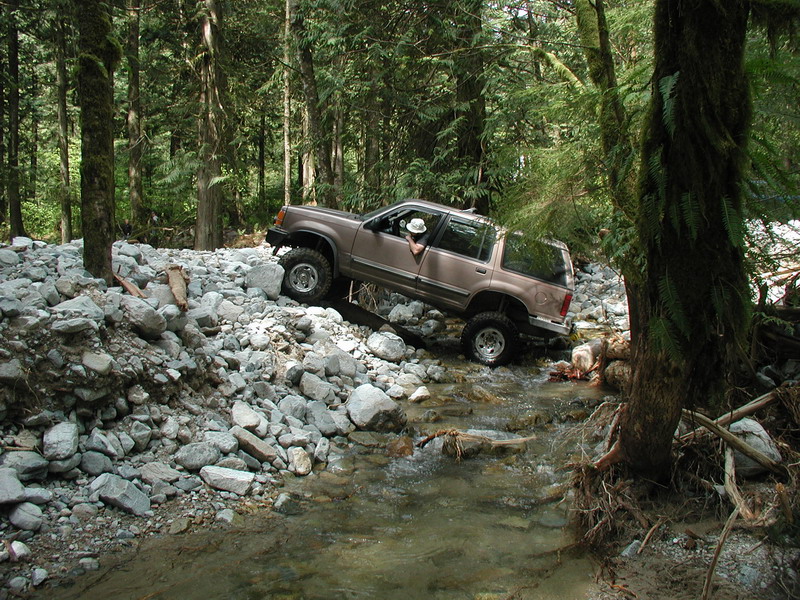 Lifted tan Ford Explorer driving through a rocky stream in a forest.
