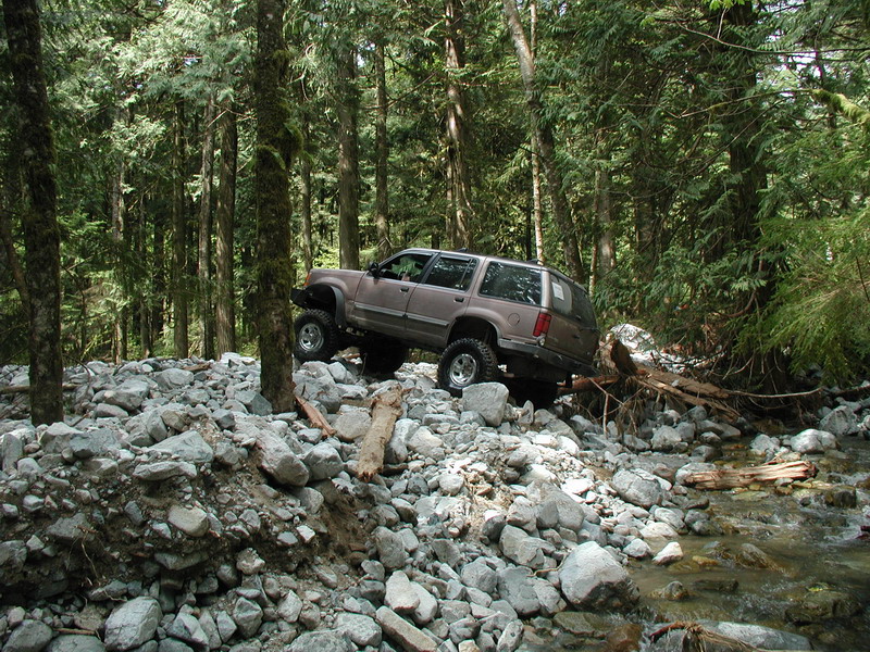 Lifted tan SUV driving over rocks in a forest next to a stream.