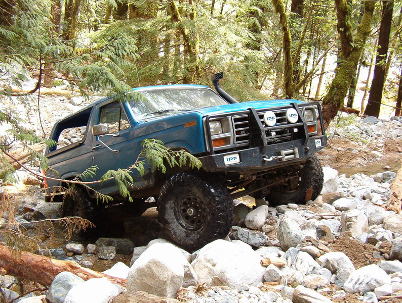 Blue Ford Bronco off-roading through a rocky stream in a forest.