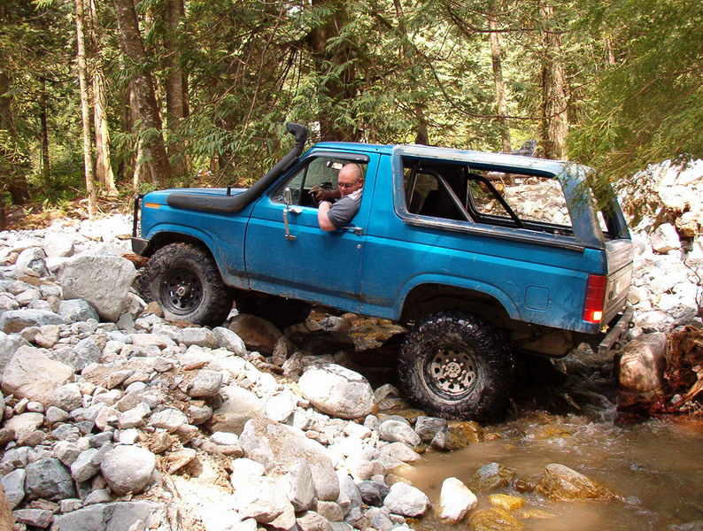 Blue 1980s Ford Bronco off-roading through a rocky stream with a snorkel.
