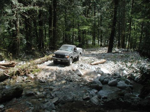 Dark pickup truck on a rocky forest trail next to a fallen log and stream.