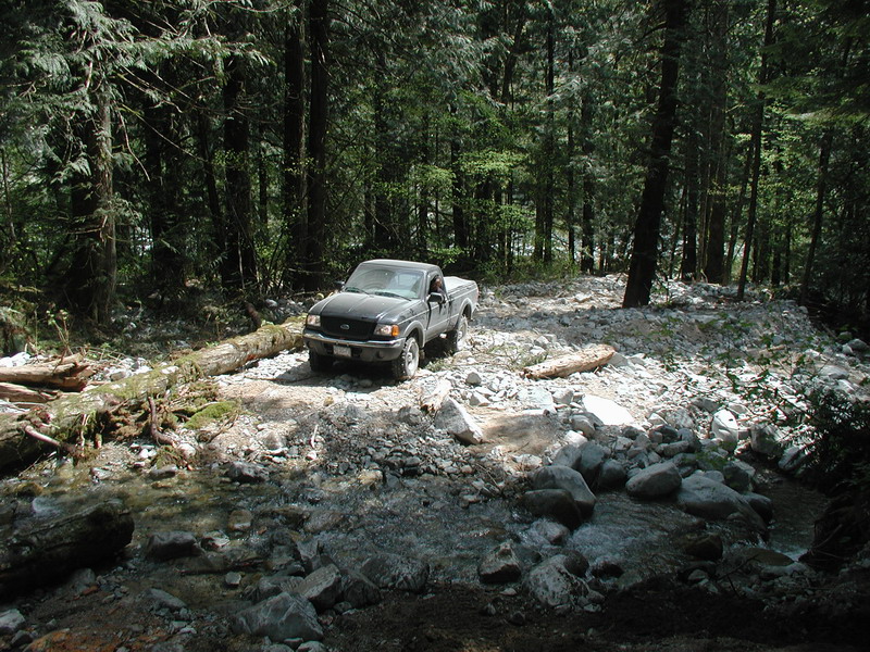 Dark pickup truck on a rocky forest trail next to a fallen log and stream.