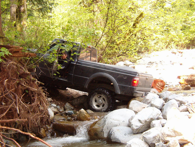 Black pickup truck driving through a rocky stream in a forest.