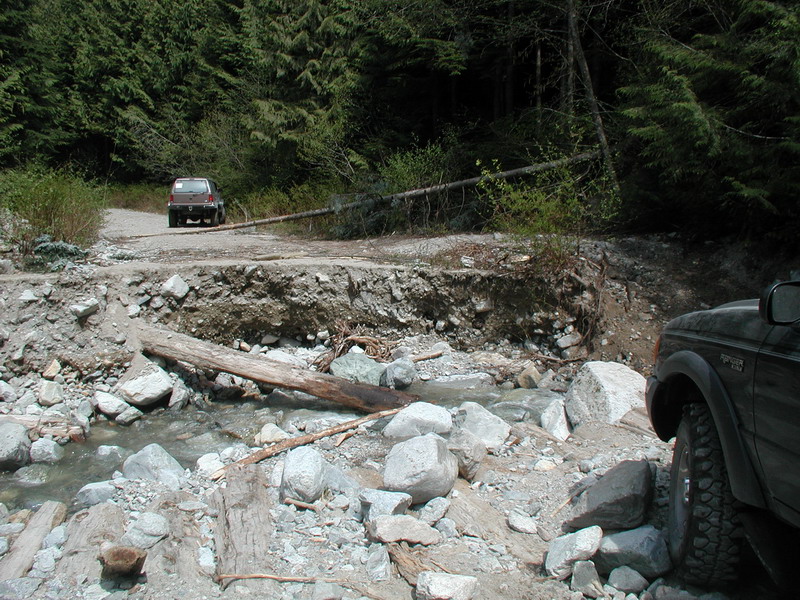 Dark SUV on a washed-out road with a stream and forest.