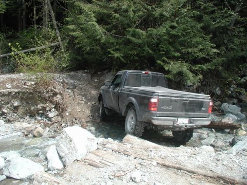 Dark Ford Ranger pickup truck driving through a rocky stream in a forest.