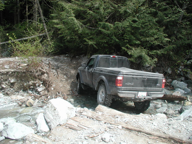 Dark Ford Ranger pickup truck driving through a rocky stream in a forest.