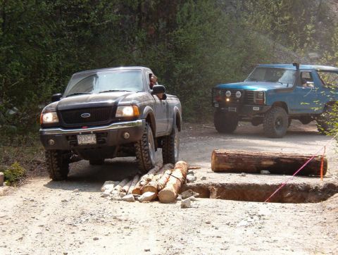A dark gray Ford Ranger pickup truck drives over logs on a dirt road, with a blue Ford Bronco behind it.