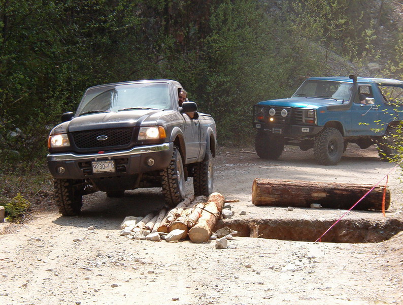 A dark gray Ford Ranger pickup truck drives over logs on a dirt road, with a blue Ford Bronco behind it.