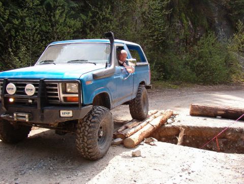 Blue Ford Bronco off-roading over logs across a trench.