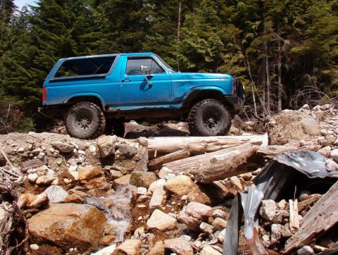 Blue 1980s Ford Bronco off-roading over logs and rocks in a forest.