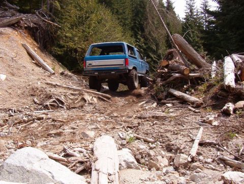 Blue 1985 Ford Bronco driving on a rough, wooded mountain trail.