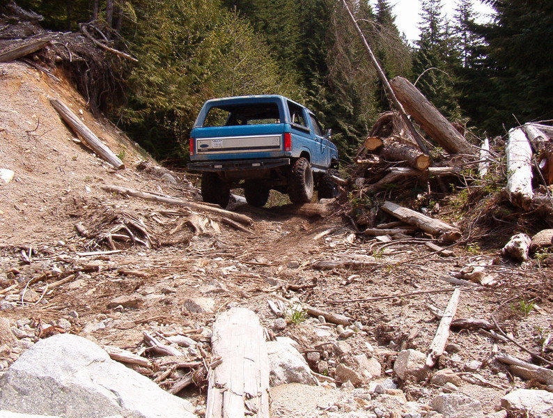 Blue 1985 Ford Bronco driving on a rough, wooded mountain trail.