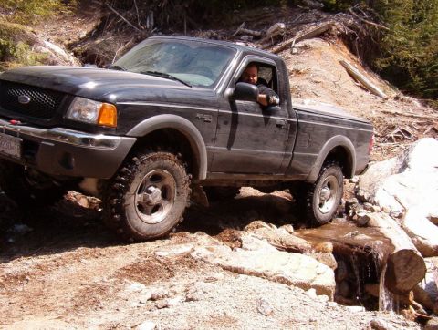 Dark pickup truck driving on a rocky off-road trail with a stream.