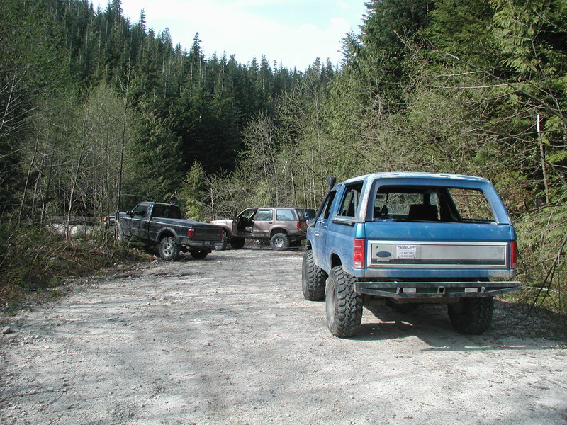 Lifted blue Ford Bronco parked on a dirt road in a forest with other vehicles.