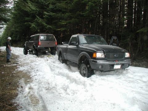 Black Ford Ranger pickup truck stuck in snow on wooded trail, being pulled by brown SUV.