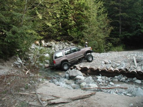 Lifted tan SUV driving through a creek over a log bridge in a forest.