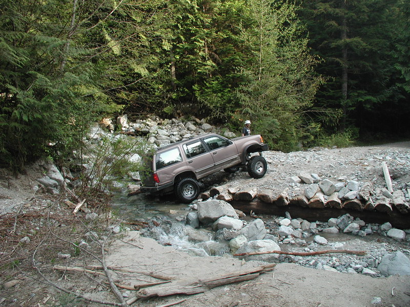 Lifted tan SUV driving through a creek over a log bridge in a forest.