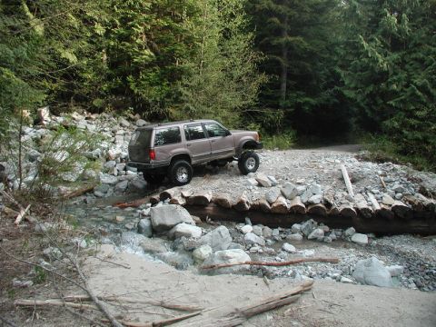 Lifted tan Ford Explorer crossing a stream on a log bridge in a forest.