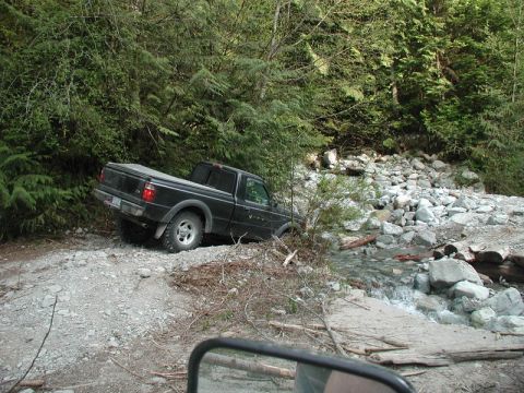 Black Ford Ranger pickup truck stuck on a rocky trail next to a stream.