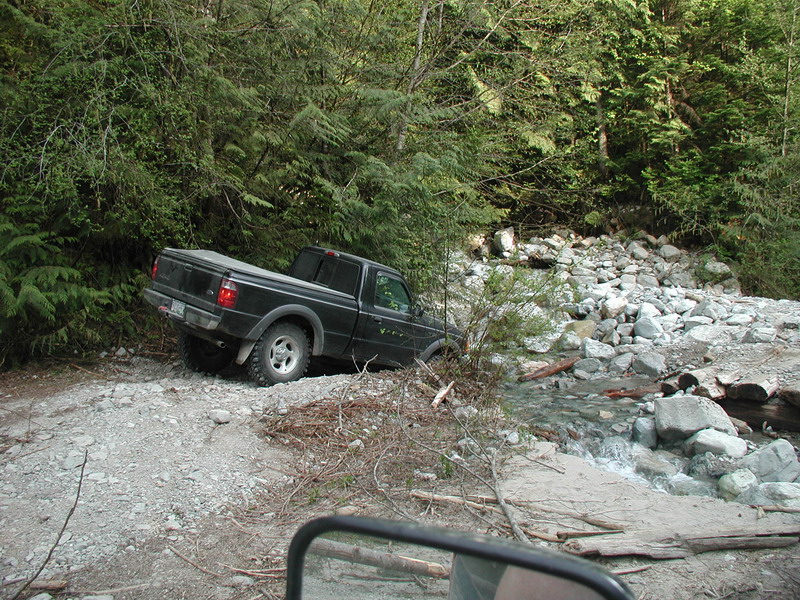 Black Ford Ranger pickup truck stuck on a rocky trail next to a stream.