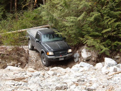 Black Ford Ranger pickup truck on a rocky off-road trail in a forest.