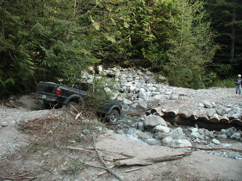 Black pickup truck stuck in a rocky stream bed with trees in the background.