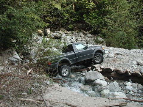 Dark pickup truck driving through a rocky stream in a forest.