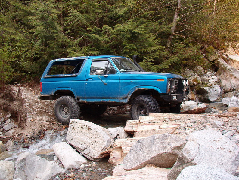 Blue 1980s Ford Bronco off-roading through a rocky stream.