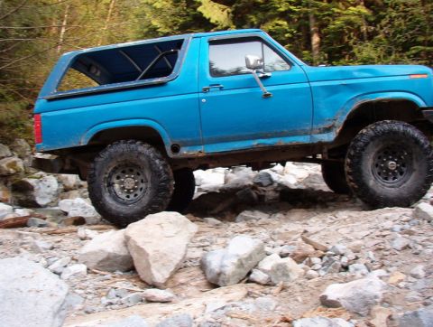 Side view of a blue 1985 Ford Bronco driving on a rocky trail.